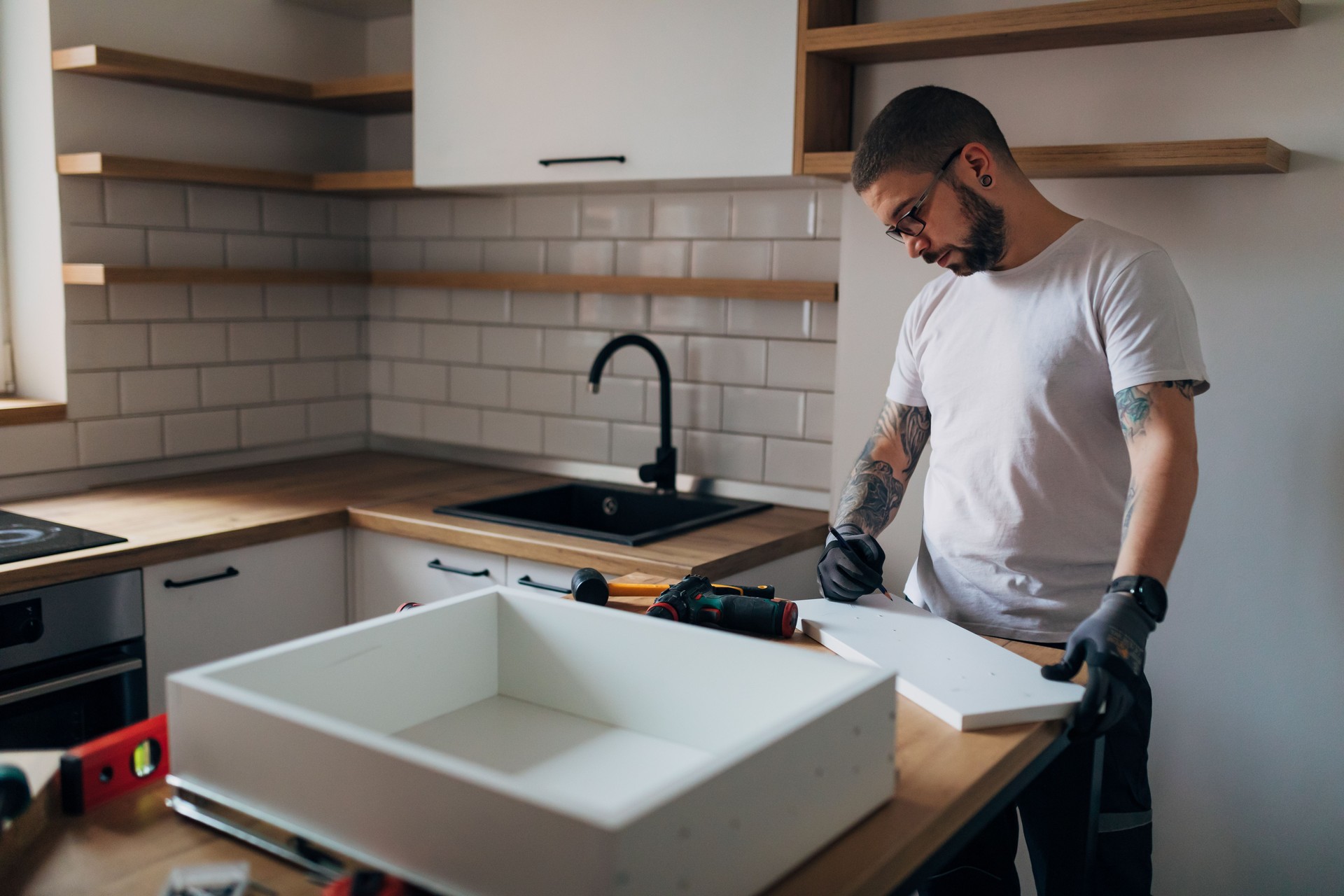 A tattooed worker is setting up a drawer in his kitchen. He is using a pencil to mark the wooden material.